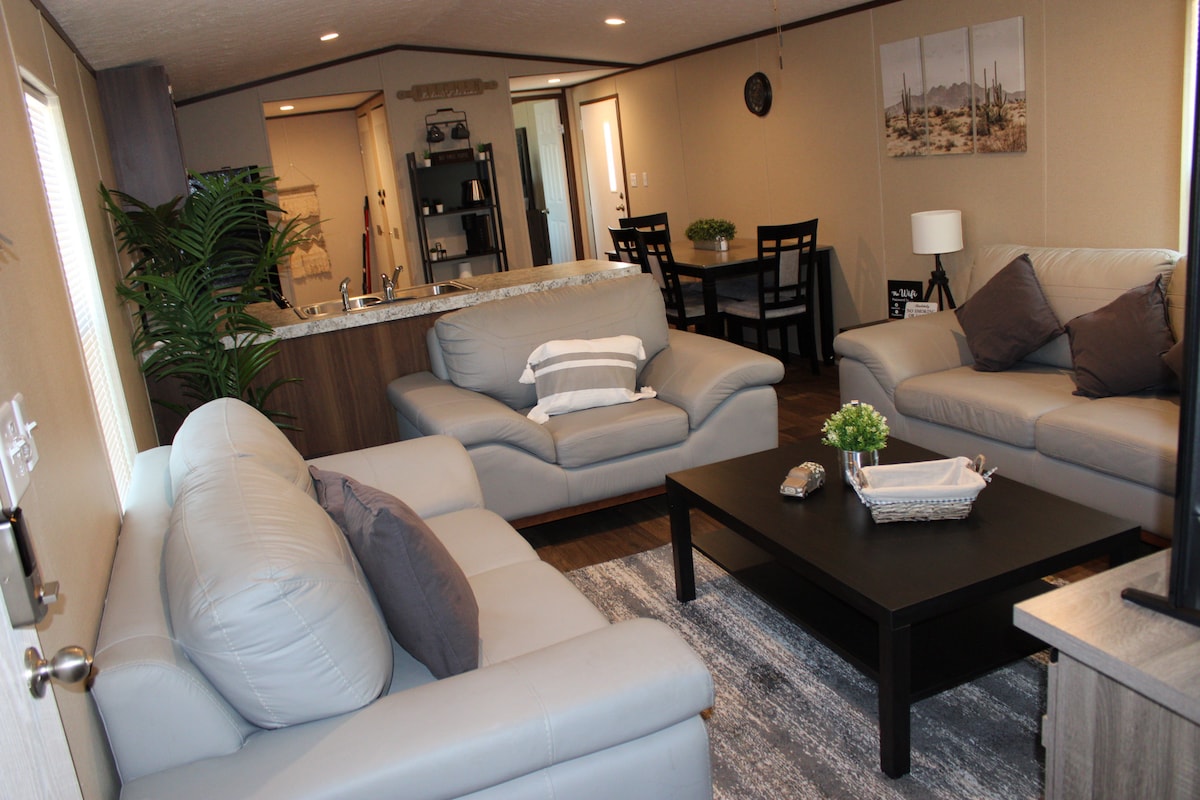 A comfortable living area features two light gray sofas and a dark coffee table placed on a textured rug. The space is open to the dining area, which includes a small table and chairs. A kitchen bar is visible in the background with modern appliances.