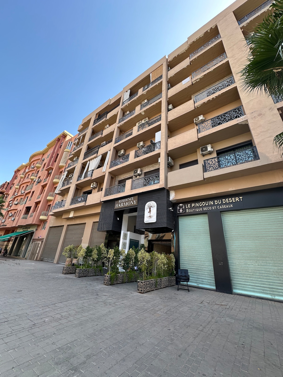 The exterior of the building showcases a modern architectural design with multiple floors. Air conditioning units are visible along the facade. Potted greenery is positioned at the entrance, providing a welcoming touch. Clear blue skies add a bright backdrop to the scene.