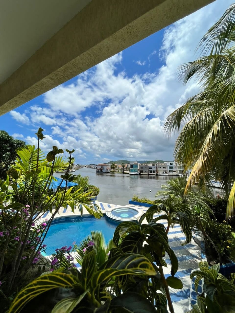 A view of the tranquil waterway is framed by lush tropical plants, showcasing a bright blue sky dotted with clouds. The inviting pool is visible below, complemented by surrounding vibrant greenery, creating a serene outdoor atmosphere.