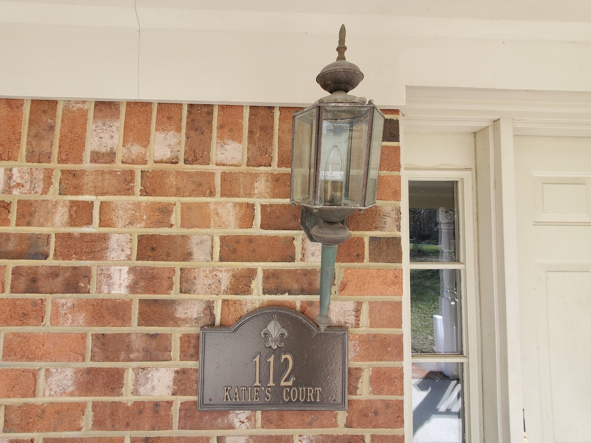 An outdoor view of a brick wall features a metal lantern mounted above a nameplate. The nameplate displays the address '112' and the title 'Katie's Court,' adorned with a decorative emblem. Natural light highlights the texture of the bricks.