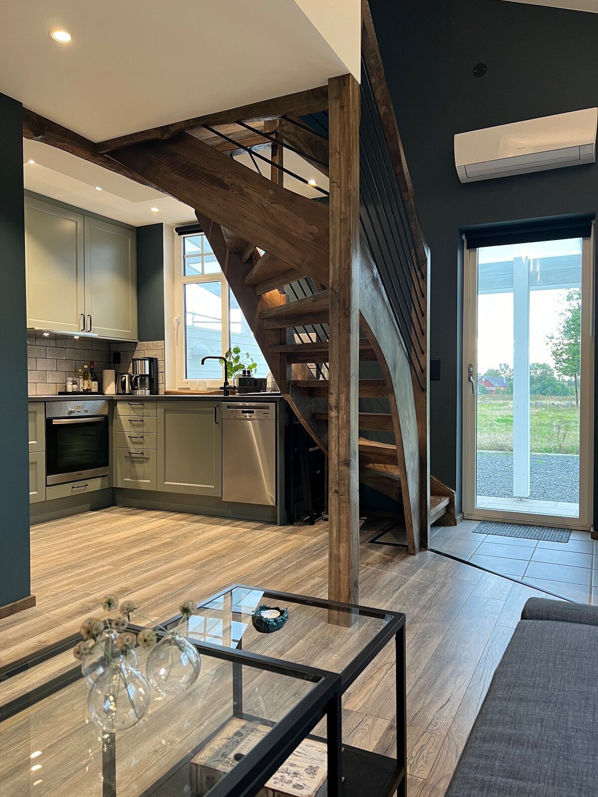 An open-plan kitchen features modern appliances and cabinetry against a dark green wall. A wooden staircase ascends to the upper level, with large windows allowing natural light and views of the outdoors. The flooring consists of light-toned tiles.