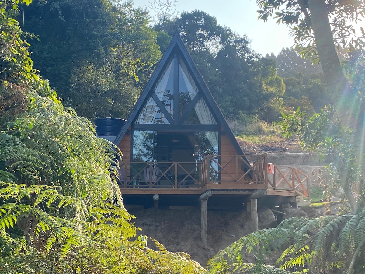 The chalet features an A-frame design with large glass panels that reflect the surrounding greenery. A wooden deck extends from the entrance, supported by stilts, providing a serene view of nature. Lush foliage frames the structure, enhancing its private and tranquil setting.