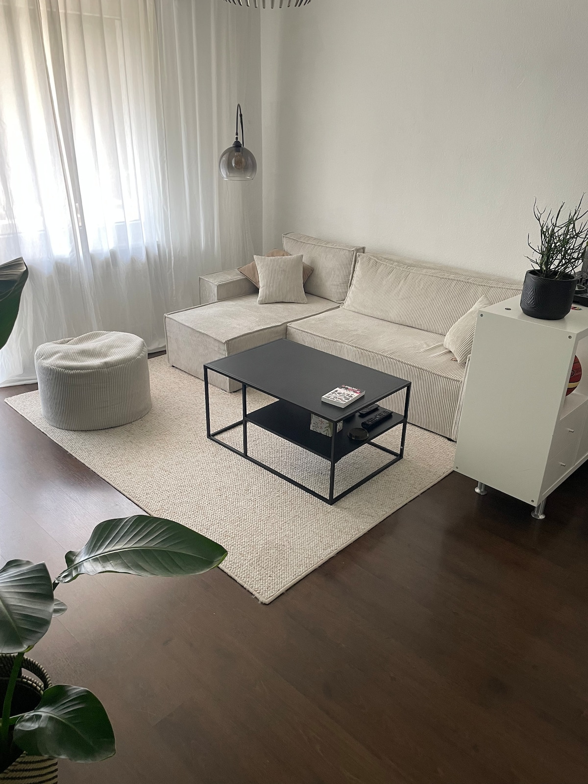 A light-filled living area contains a beige sofa and a round pouf, both positioned on a neutral-colored area rug. A black coffee table sits in the center, adorned with a small stack of magazines. Soft curtains gently diffuse natural light from the window.