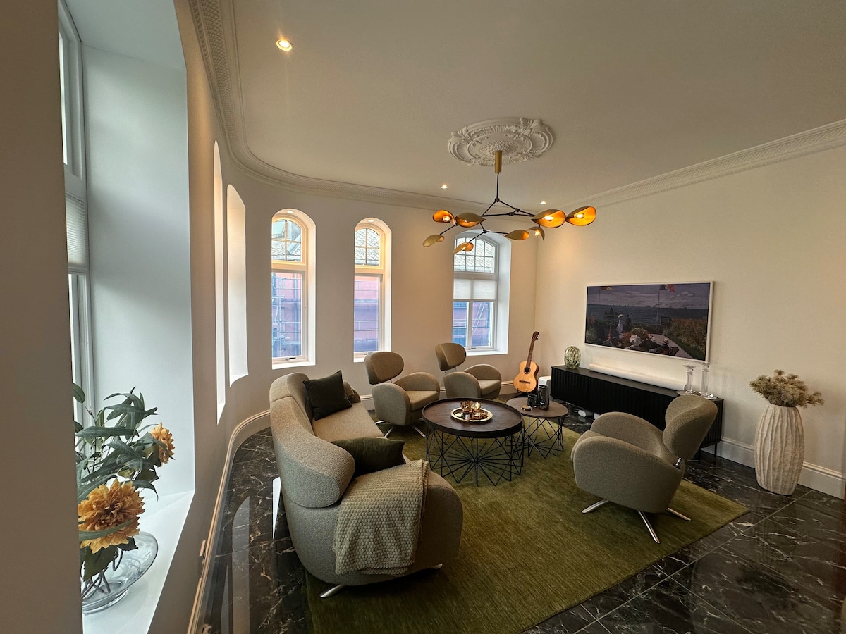 A spacious living area showcases an arrangement of curved, upholstered chairs surrounding a low coffee table on a green area rug. A large television is positioned on a dark cabinet, while sunlight filters through arched windows, highlighting a guitar and decorative plants.