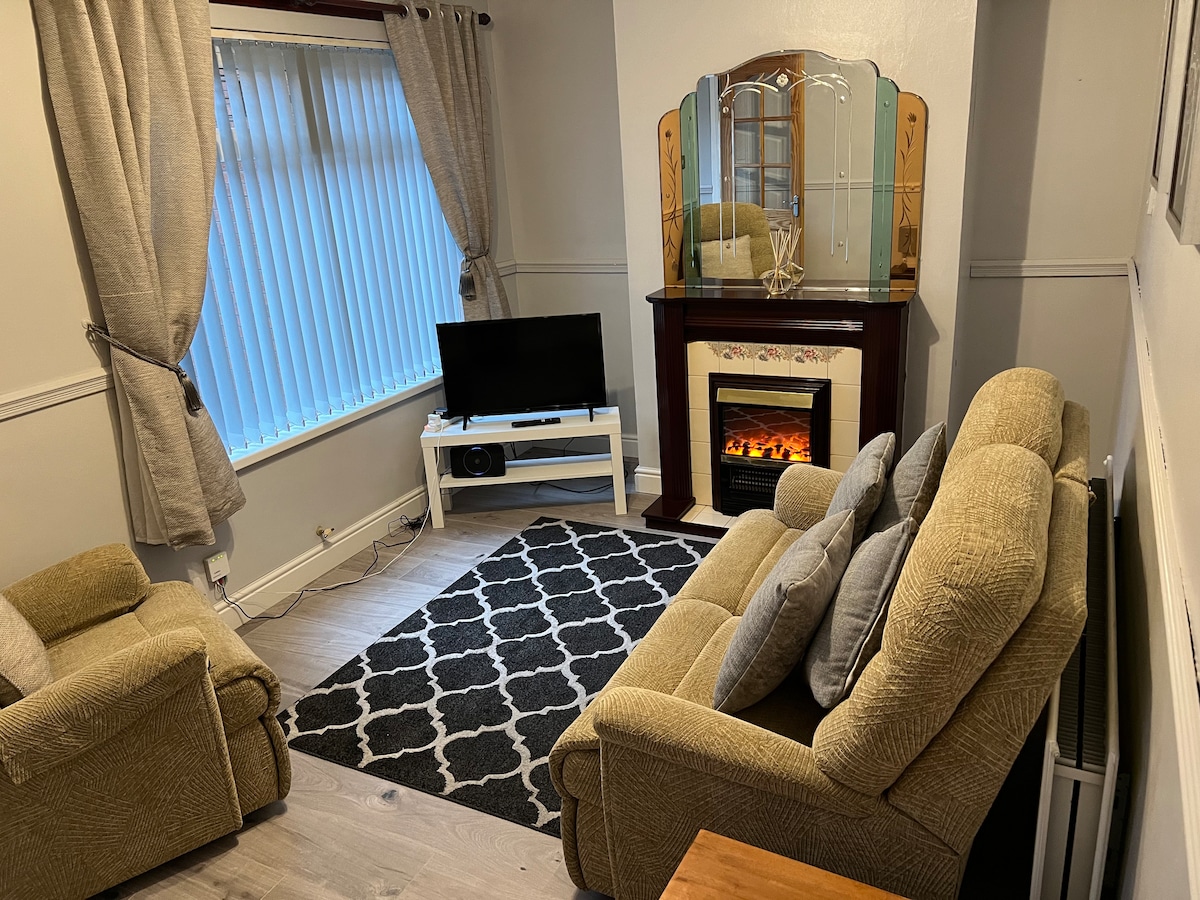A small living area features a cozy seating arrangement with two armchairs and a sofa adorned with cushions. A television is positioned on a low stand, opposite a decorative fireplace. Soft light enters through a window covered by vertical blinds, while a patterned rug adds warmth to the hardwood floor.
