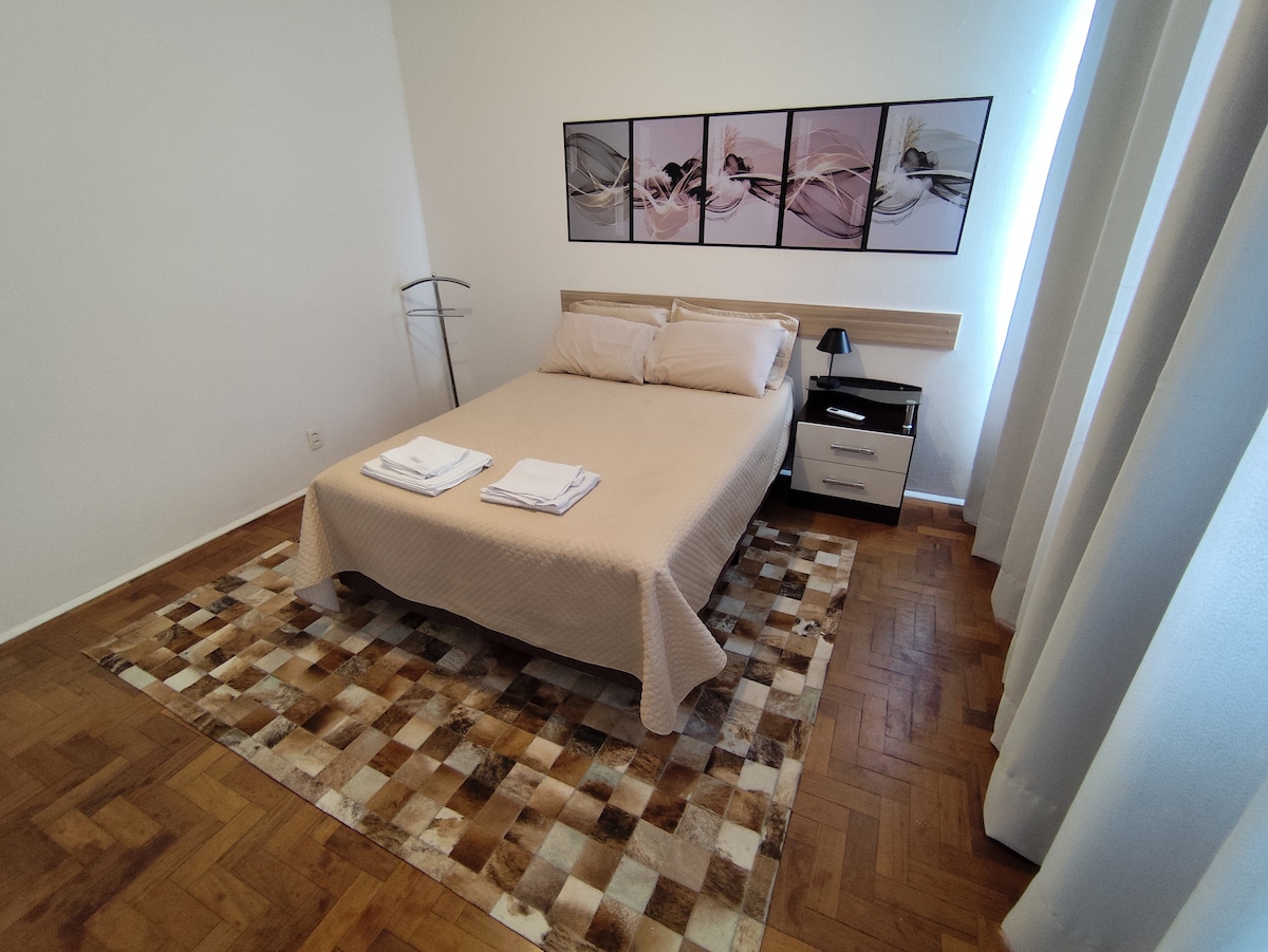 A spacious bedroom features a comfortable bed with light linens, accompanied by a nightstand and lamp. Elegant wall art enhances the room's decor, while a patterned rug adds texture. Natural light filters in through sheer curtains, creating a bright and welcoming ambiance.