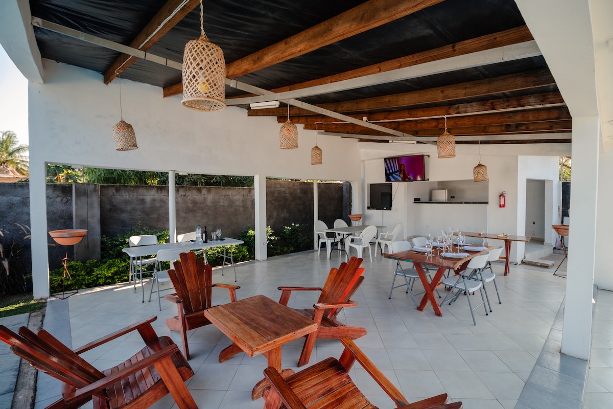 An open outdoor seating area features wooden tables and chairs, arranged to encourage relaxation and conversation. Decorative woven light fixtures hang from the ceiling, providing a warm ambiance. The space is complemented by greenery visible in the background and simple, modern decor.