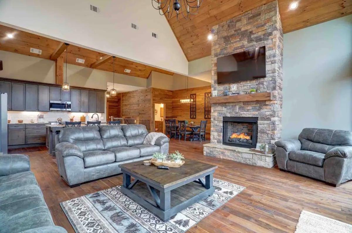 A spacious living area features dark leather sofas arranged around a wooden coffee table. A stone fireplace is set into the wall, providing a focal point. The high ceiling is complemented by wooden beams, and a large television hangs above the fireplace. The adjoining kitchen is visible in the background.