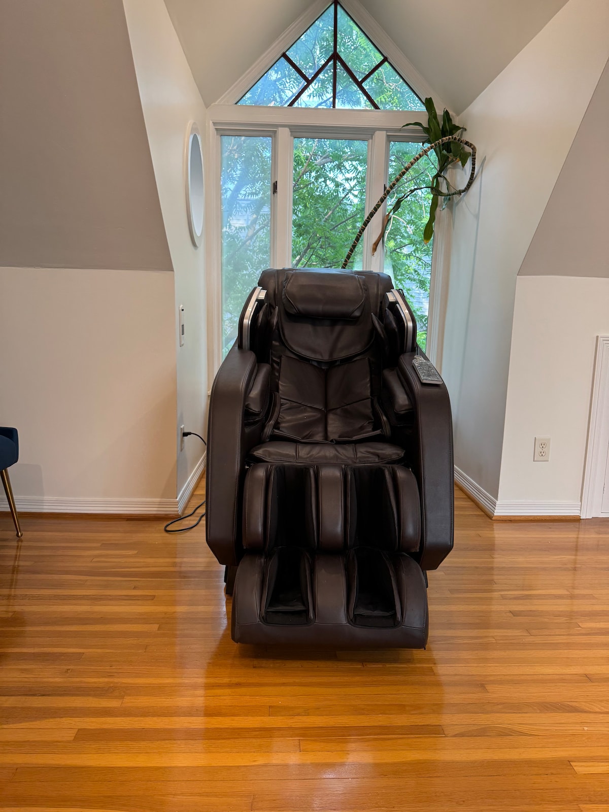 A massage chair is positioned in a well-lit corner, featuring sleek black upholstery. The chair is surrounded by large windows that allow natural light to brighten the room, and a green plant adds a touch of nature to the space.