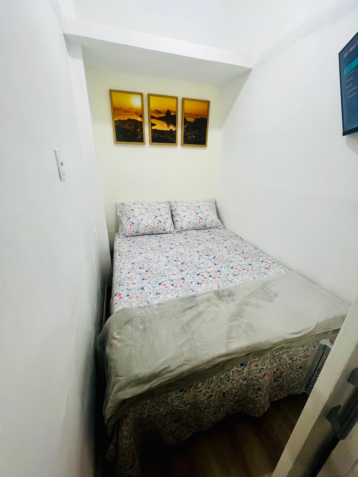 A compact sleeping area is seen featuring a spacious bed dressed in a floral-patterned coverlet. Three framed artworks hang above the bed, while a smooth white wall provides a clean backdrop. Light wood flooring adds warmth to the space.