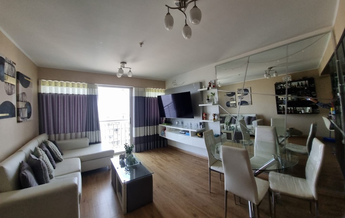 A living area is highlighted by a large window, allowing natural light to fill the space. A comfortable white sofa faces a wall-mounted television. A glass coffee table sits in the center of the room, surrounded by modern dining chairs, while mirrors enhance the sense of openness.
