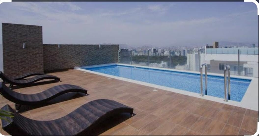 A rooftop terrace features a shimmering pool bordered by stylish lounge chairs, offering expansive views of the city skyline. The pool area is illuminated by soft natural light, with a clear sky overhead and a mixture of urban architecture in the background.