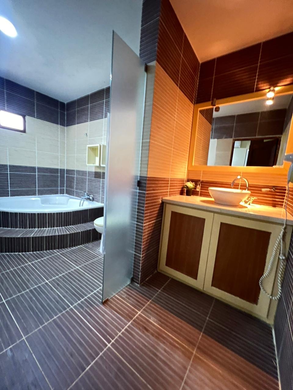 A modern bathroom features a spacious corner bathtub alongside a practical sink area with wooden cabinetry. The walls are tiled in a contrasting pattern of light and dark hues, while natural light enters from a small window above, enhancing the overall brightness of the space.