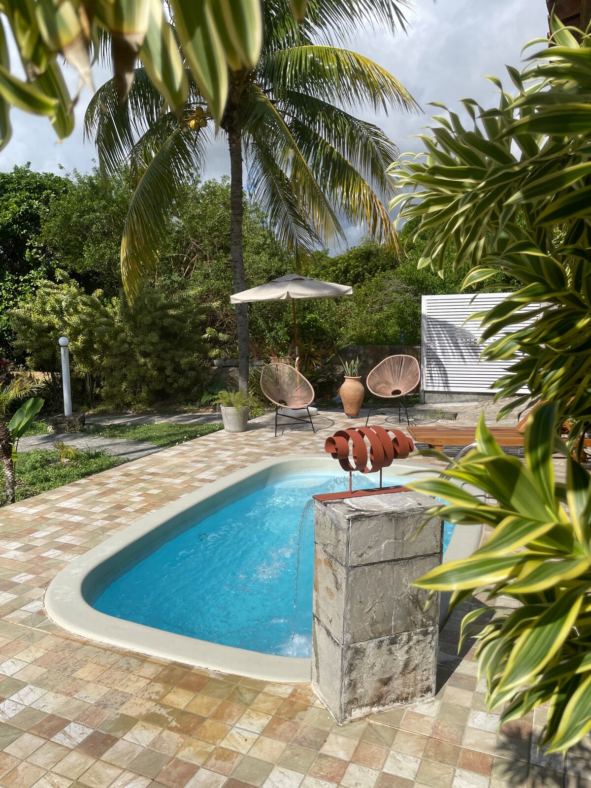 A serene outdoor space showcases a compact swimming pool surrounded by tropical plants. Two stylish lounge chairs are positioned under a shaded umbrella, while a whimsical sculpture adds a touch of charm. A palm tree partially frames the scene, enhancing the natural feel.