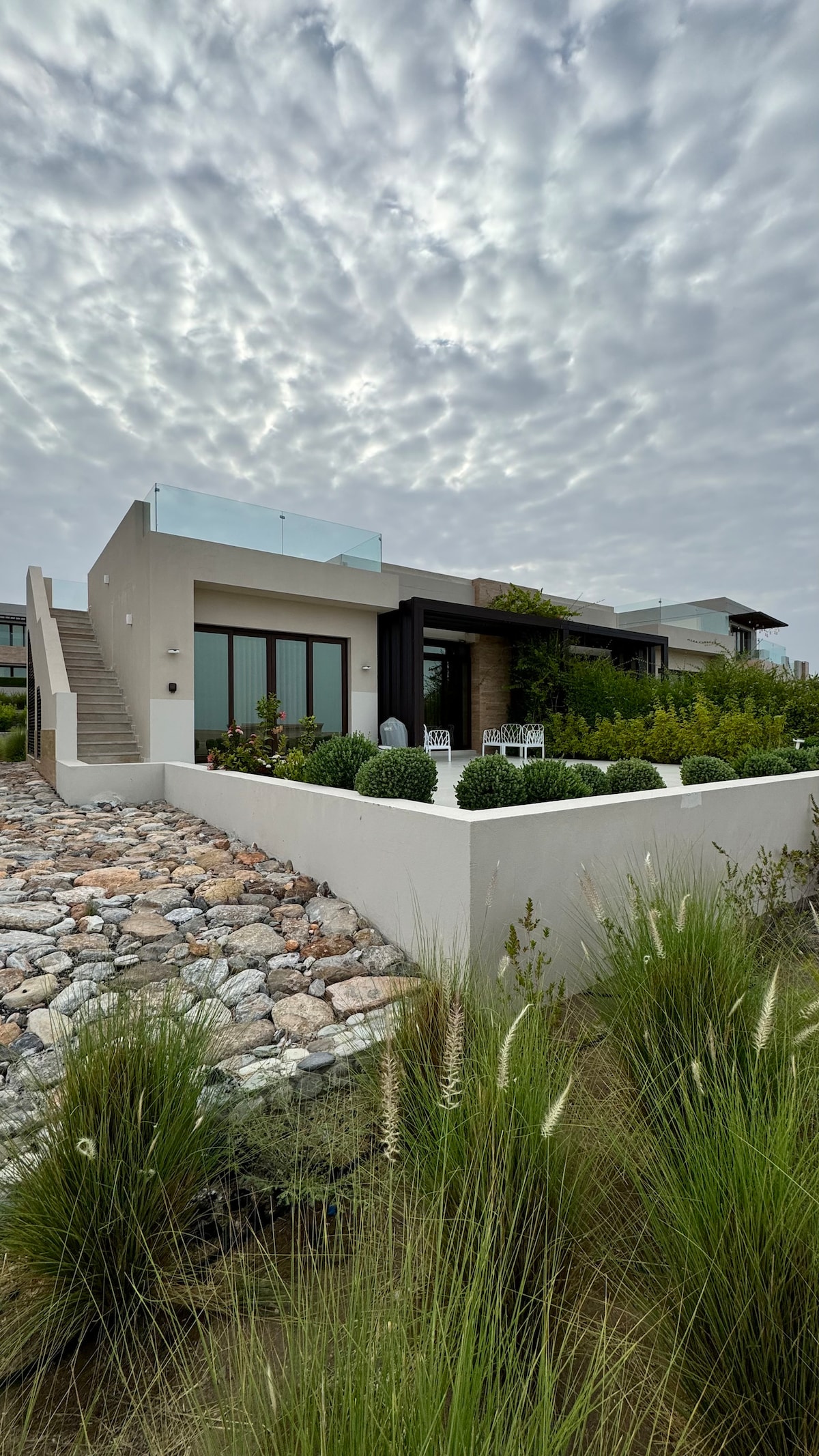 A modern house is displayed with a landscaped garden featuring ornamental grasses. Smooth stone pathways lead to the entrance. A terrace with glass railings is visible above, and large windows invite natural light into the living spaces. The sky is overcast, adding to the tranquil setting.