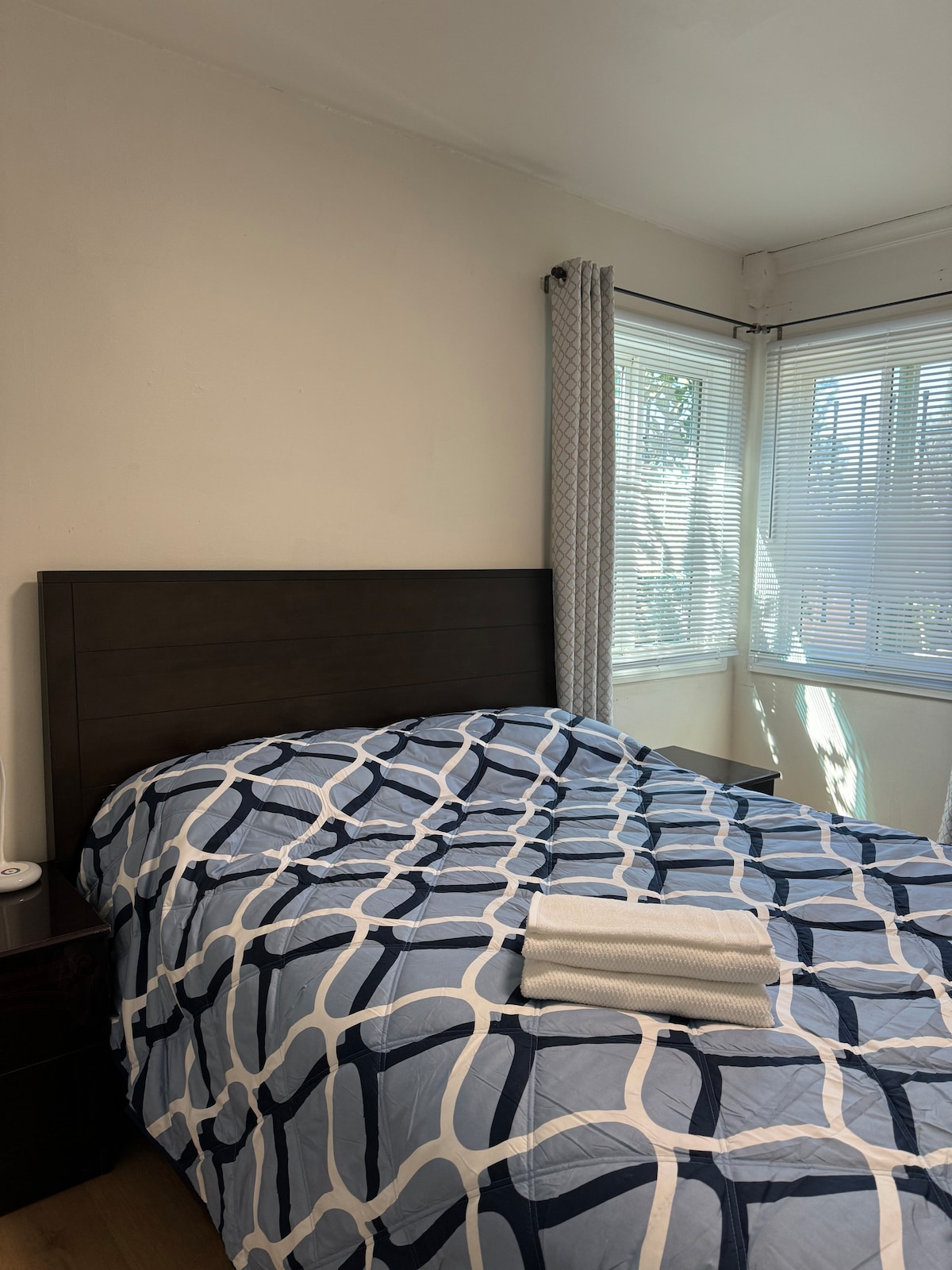 A private room features a spacious bed dressed in a blue and white patterned comforter. Natural light filters through multiple windows with light curtains, illuminating the room. A bedside table holds a small lamp, while neatly folded towels are placed on the bed.