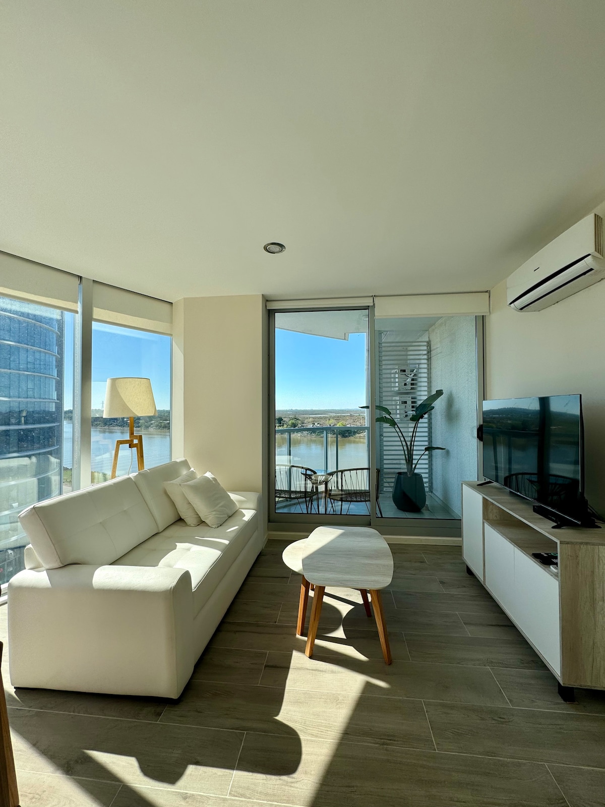 A bright living area features a white sofa complemented by decorative pillows. A small table sits in front, with a television mounted on the wall. Large windows allow natural light to fill the space, revealing a view of the outdoors and greenery.
