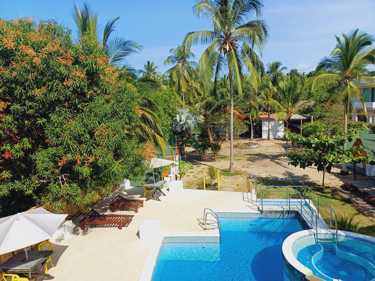 A sunlit outdoor area features a swimming pool surrounded by lounge chairs and umbrellas. Lush palm trees provide shade and greenery, while a nearby dining area with wooden tables enhances the inviting space. The scenery blends natural elements with recreational amenities.