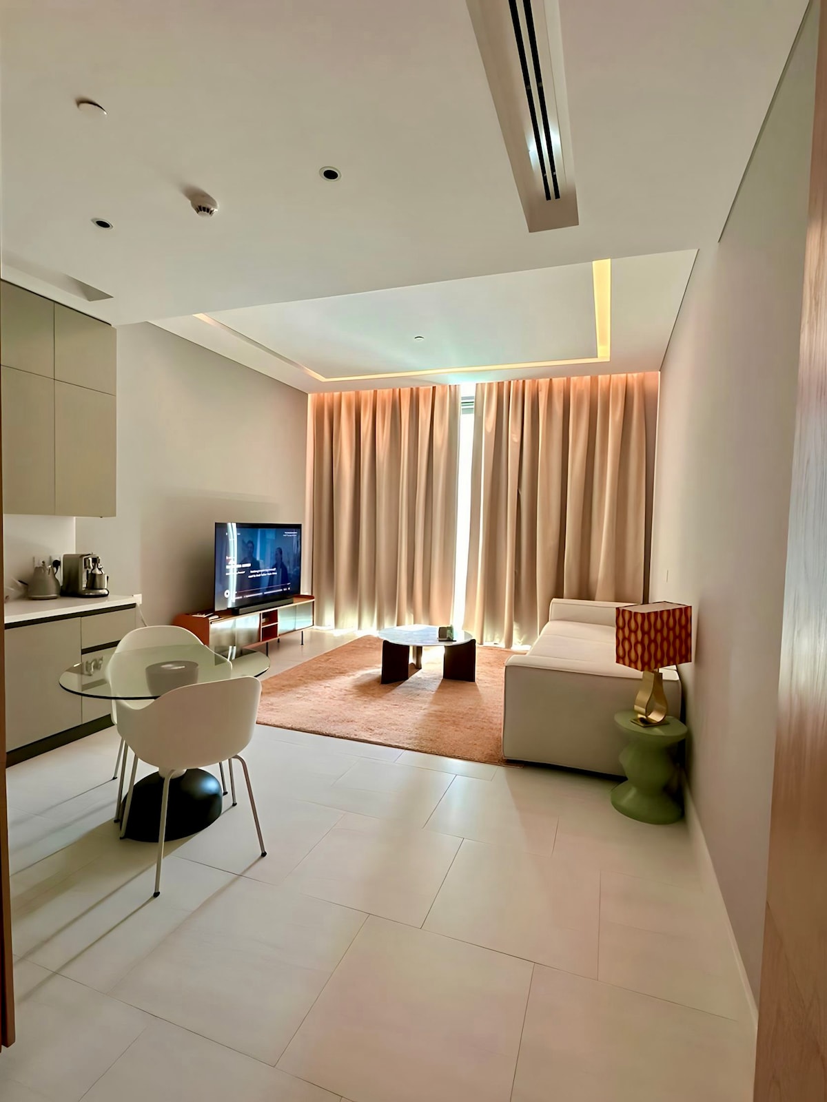 A modern living area features a comfortable sofa beside a contemporary coffee table. A TV stands against the wall. A dining table with seating for two is positioned next to a kitchenette, with light beige walls and soft natural light filtering through floor-to-ceiling curtains.