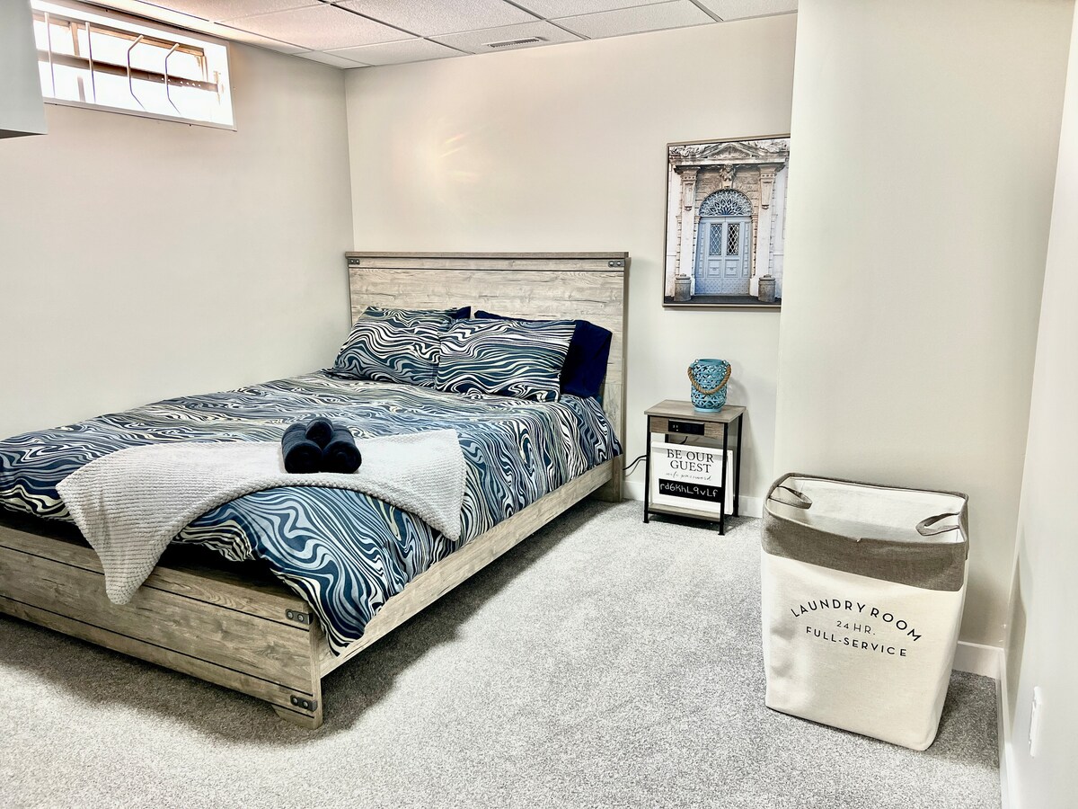 A spacious sleeping area features a comfortable bed with a patterned blue and white duvet cover. Soft grey carpeting extends across the floor. A laundry basket is positioned next to a bedside table, which holds a decorative bowl and framed artwork of a doorway.