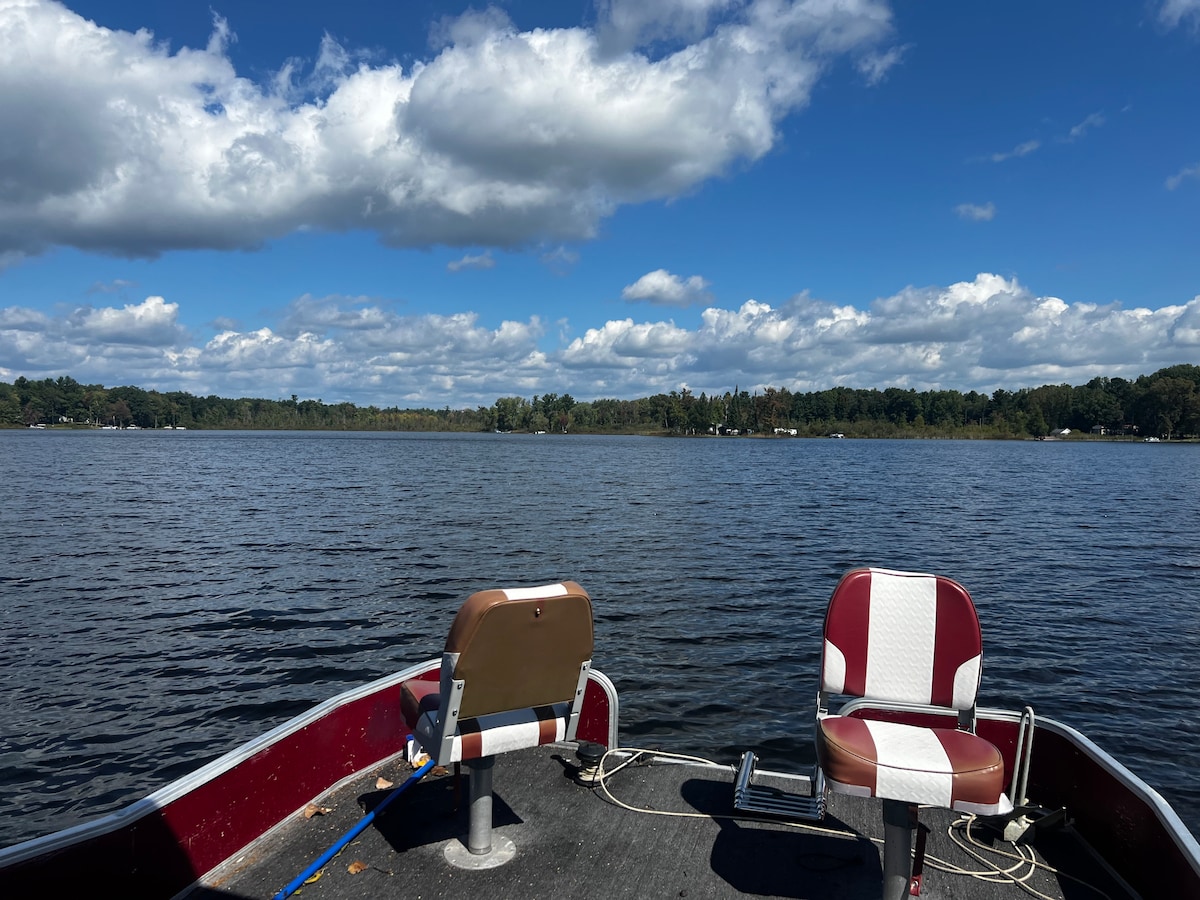 A calm lake scene is captured from a boat, showcasing a broad expanse of water under a partly cloudy sky. Two padded boat seats are visible at the forefront, indicating comfort for passengers. Lush green trees line the distant shoreline, enhancing the serene atmosphere.