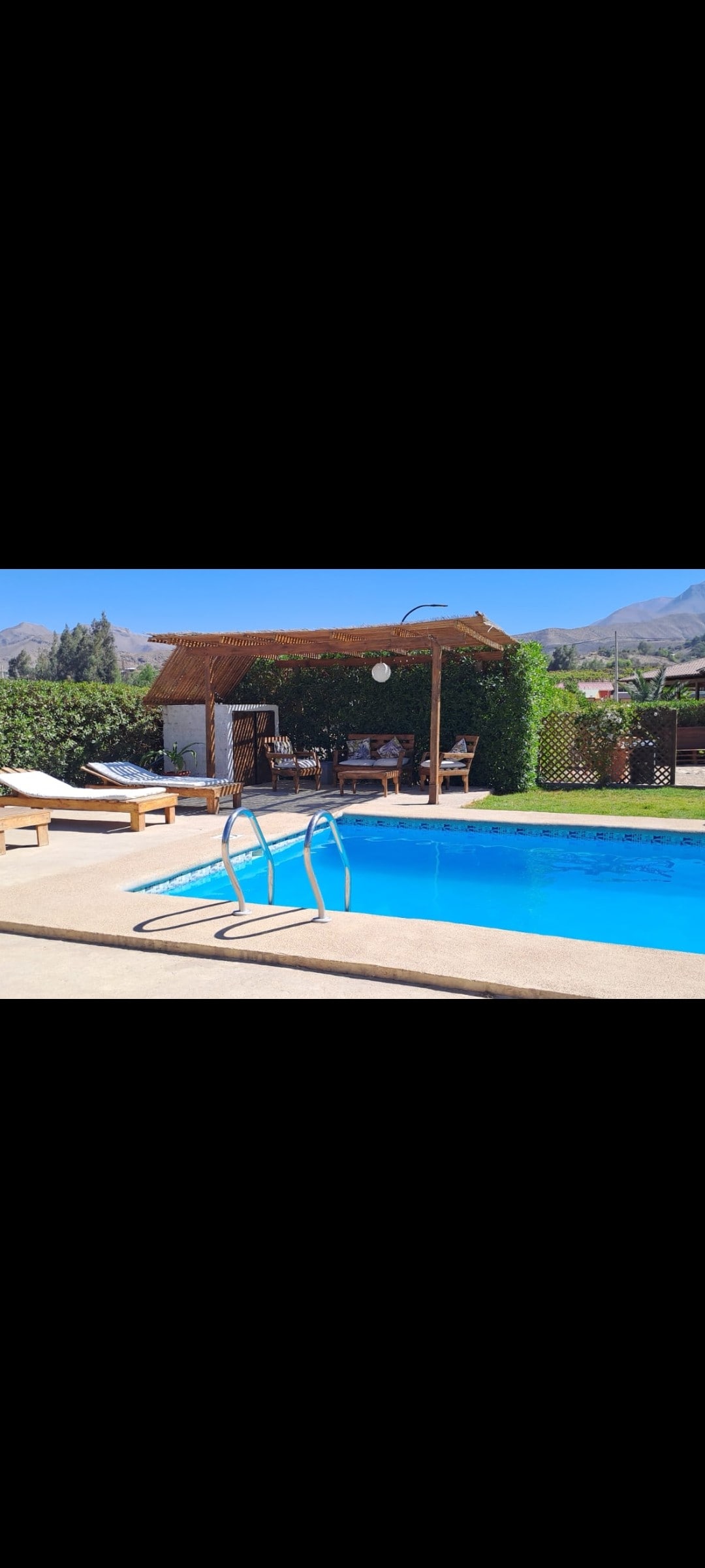 A sunlit pool area is present, featuring a refreshing blue swimming pool with a ladder leading into the water. Comfortable loungers are set beside the pool, and a thatched-roof structure provides shaded seating, surrounded by lush greenery and mountain views.