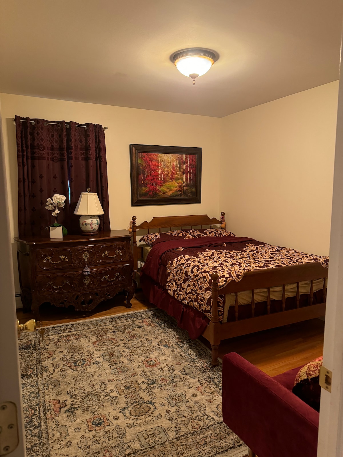 A neat bedroom is shown, featuring a wooden bed covered with rich, patterned bedding. A painting of a forest hangs above the bed, complemented by a bedside lamp. An ornate dresser and a chair add to the room's furnishings, with warm lighting provided by a ceiling fixture.