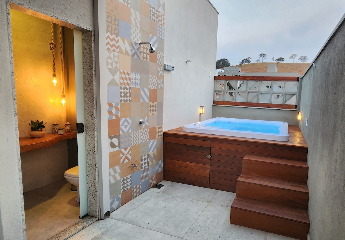 An outdoor area features a wooden hot tub surrounded by contemporary tiles and a textured wall. Soft lighting is provided by wall sconces, and a small plant is placed on a wooden countertop. A nearby restroom is discreetly located behind a partition.