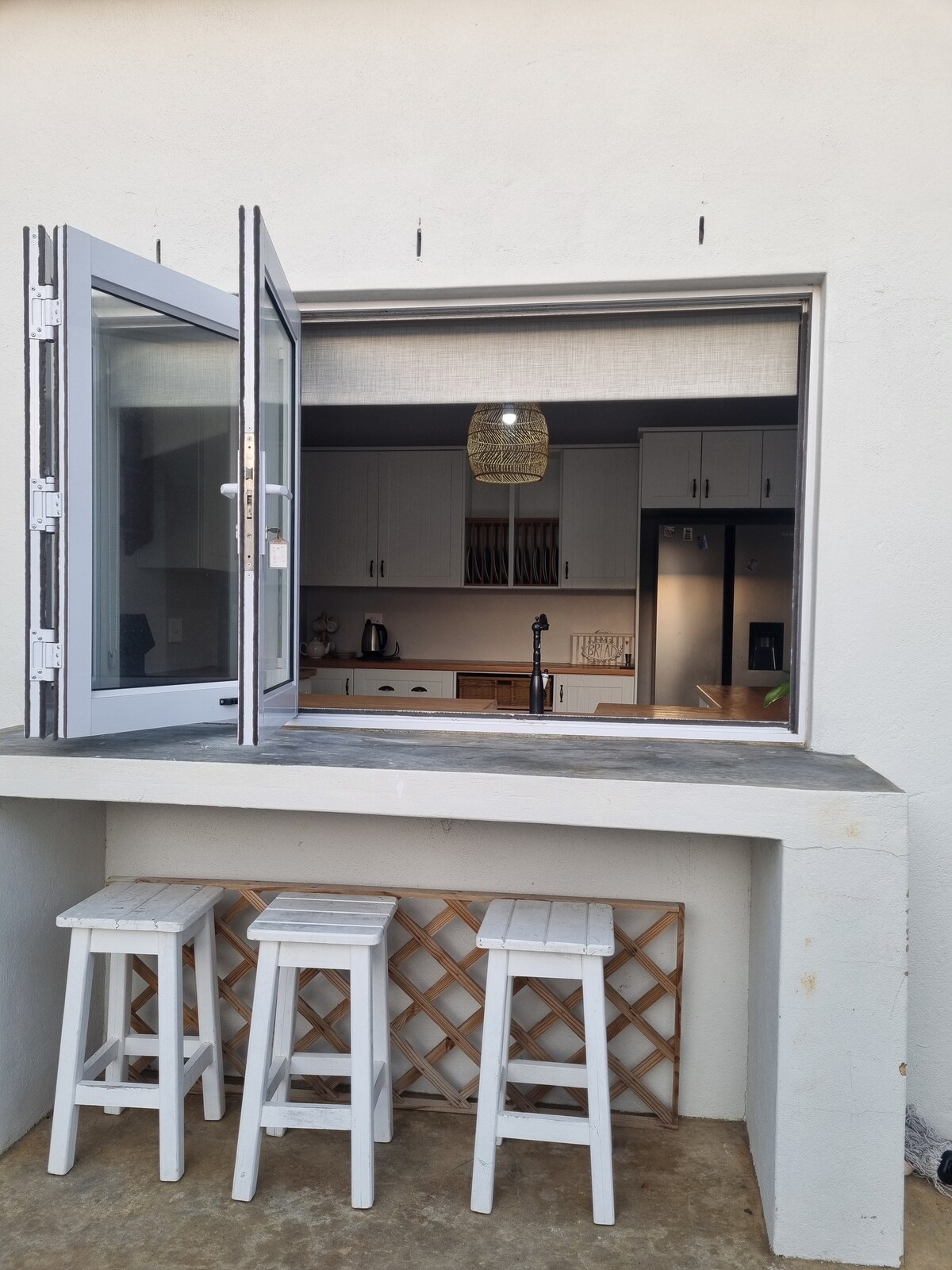 An exterior view reveals a kitchen window with two panels opened wide, displaying a modern kitchen setup inside. A stone countertop is complemented by three white bar stools positioned beneath the window, providing a casual seating area for guests.