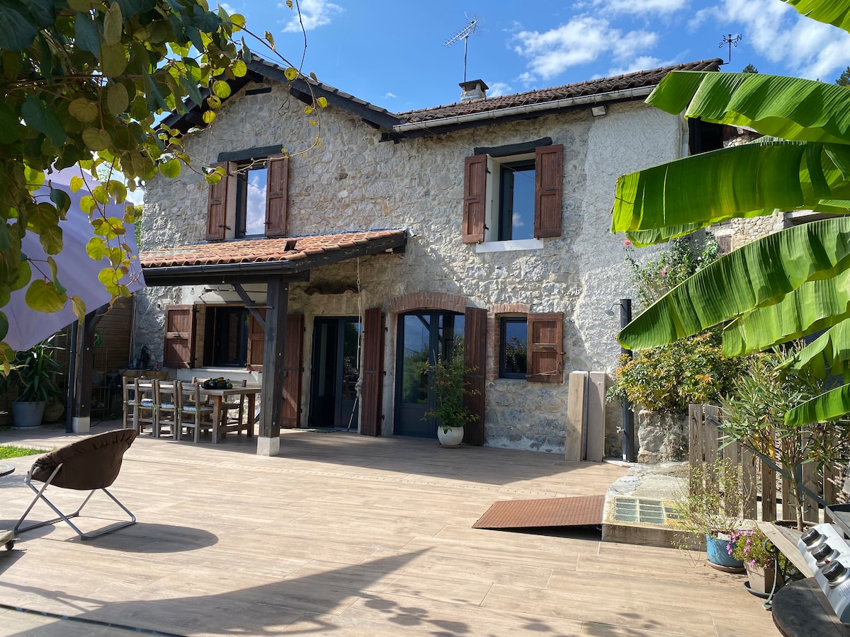 A charming stone house is set against a clear blue sky, featuring wooden shutters and a covered patio area. Outdoor seating is arranged on a spacious deck, complemented by greenery and potted plants, creating an inviting outdoor space.