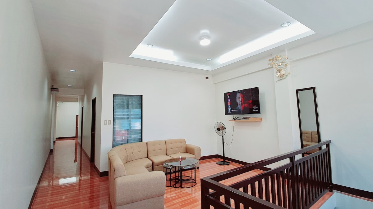 A spacious living area features a light-colored sectional sofa and a round coffee table. A large wall-mounted television is visible, along with a fan positioned nearby. The room is illuminated by recessed lighting, creating a bright and welcoming environment.