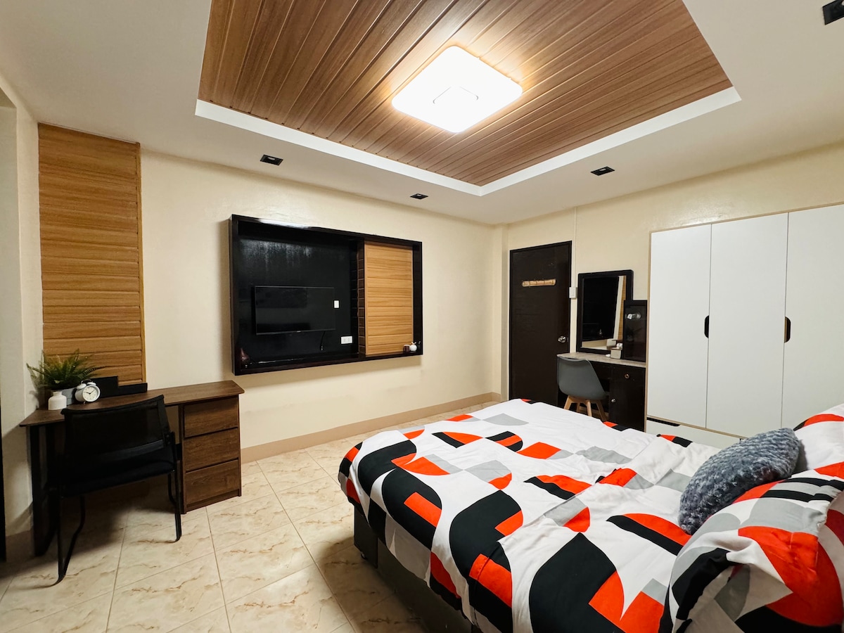 A spacious bedroom features a modern design with a color-blocked bedding set. The room includes a flat-screen TV mounted on the wall, a workstation with a chair, and a wardrobe with sliding doors. Soft lighting is provided by a ceiling fixture, enhancing the overall ambiance.