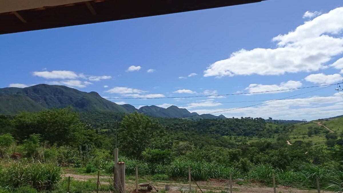 A sweeping view of rolling hills and lush green landscapes is captured beneath a clear blue sky, dotted with soft clouds. The foreground features a mix of trees and vegetation, enhancing the natural beauty of the surrounding area.
