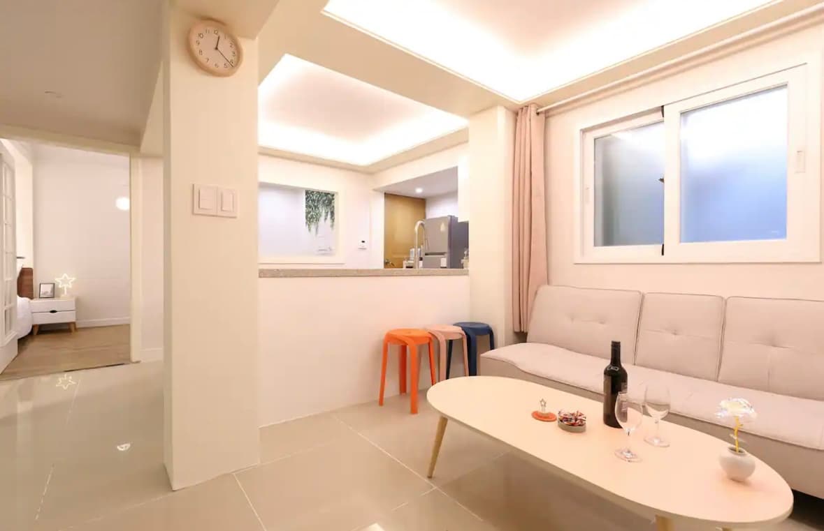 A well-lit living area showcases a neutral-toned sofa paired with bright orange and blue stools. A round coffee table sits centrally, adorned with glasses and a bottle. Soft curtains frame the window, allowing natural light to enhance the inviting space.