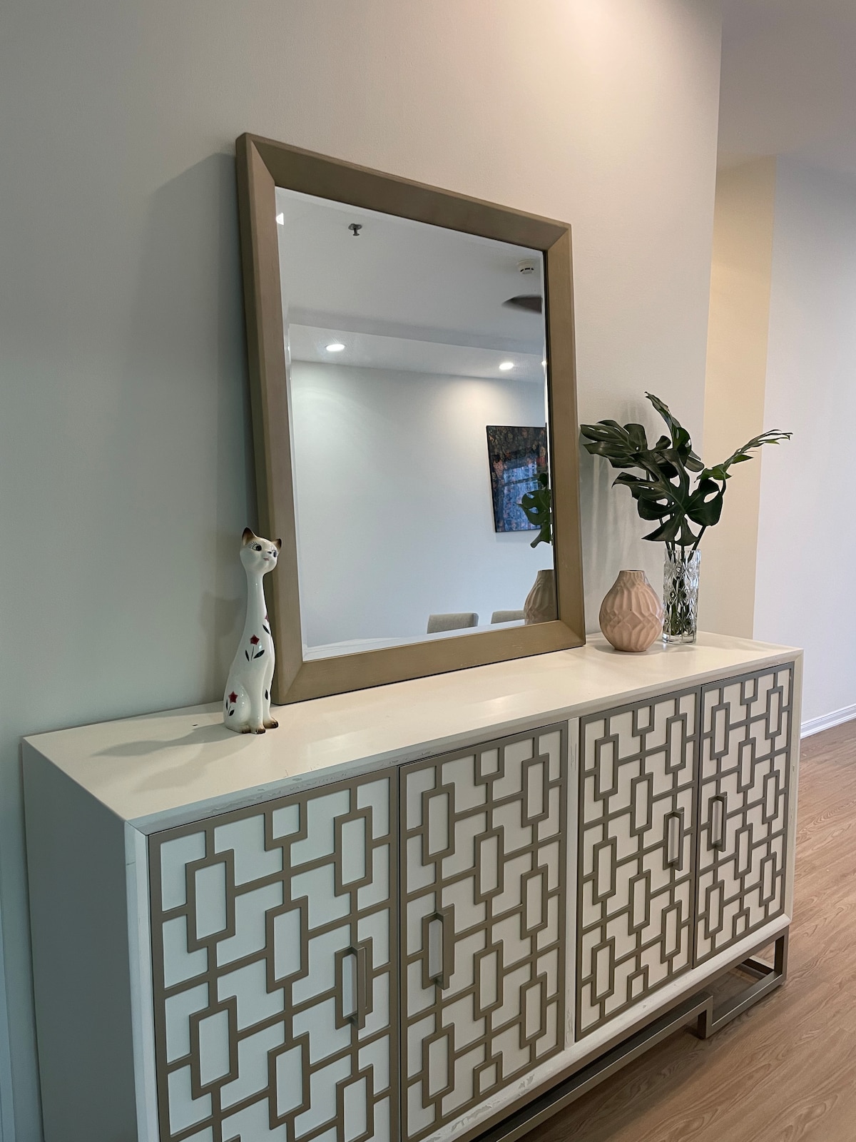 A modern console table is positioned against a light-colored wall, featuring decorative cabinetry with geometric patterns. A large, framed mirror hangs above it, reflecting the space. A slender vase with green foliage and a sculptural piece are placed on the table, adding subtle decor elements.