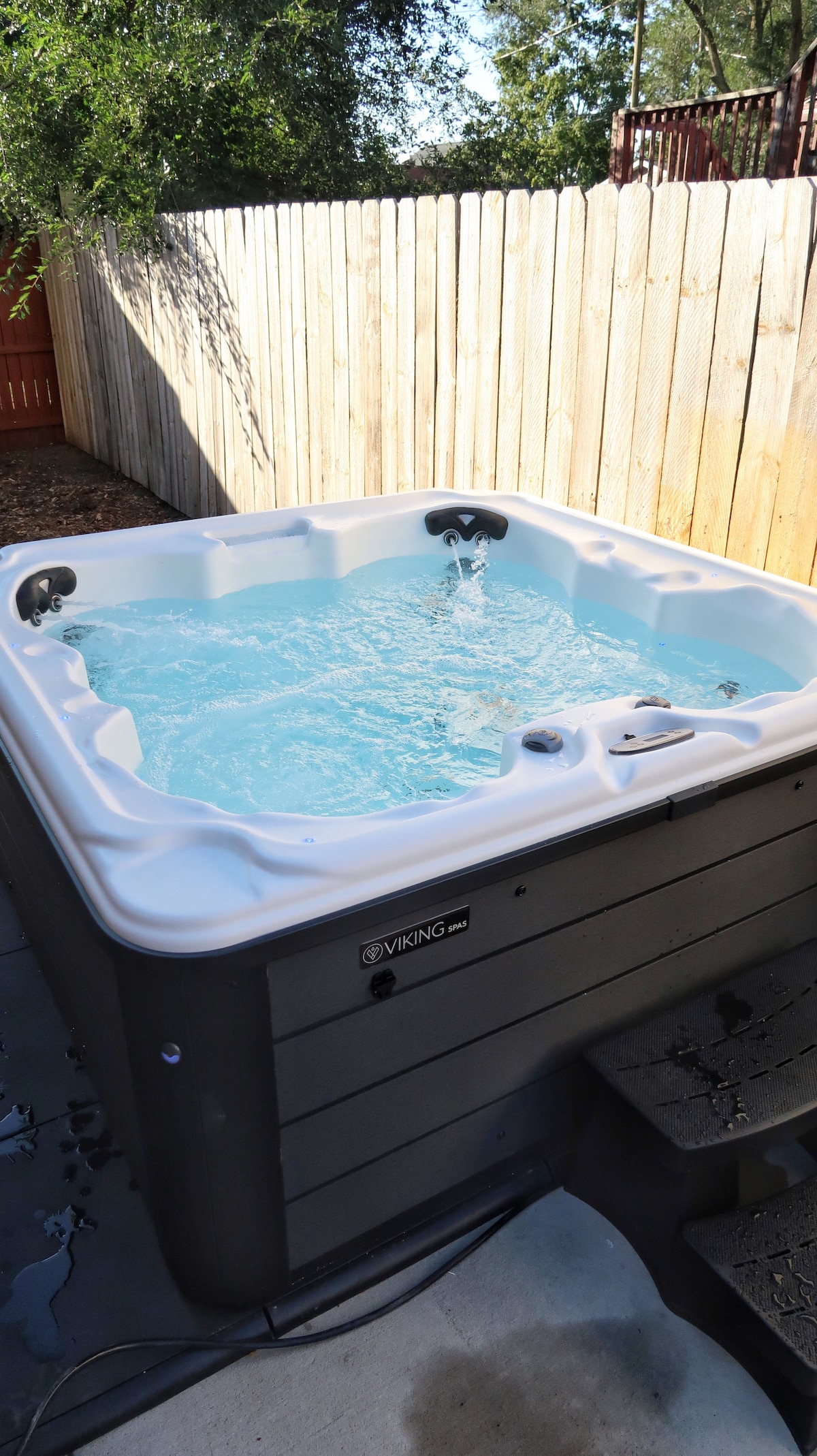 A hot tub is visible in a private outdoor area, featuring a smooth, white surface with water gently bubbling. Surrounding the hot tub, a wooden fence provides privacy, while a concrete surface leads to the steps for easy access.