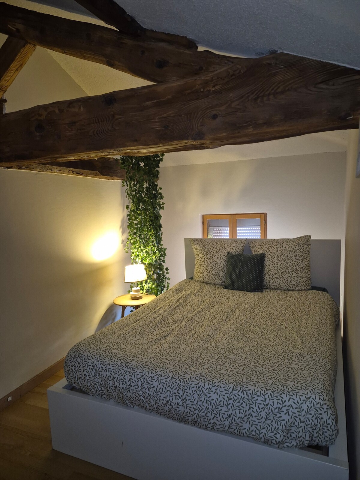 A cozy sleeping area features a spacious bed with neutral-toned bedding. Natural wood beams are visible overhead, adding texture to the space. A small side table with a lamp sits next to the bed, alongside a potted plant that introduces a touch of greenery.