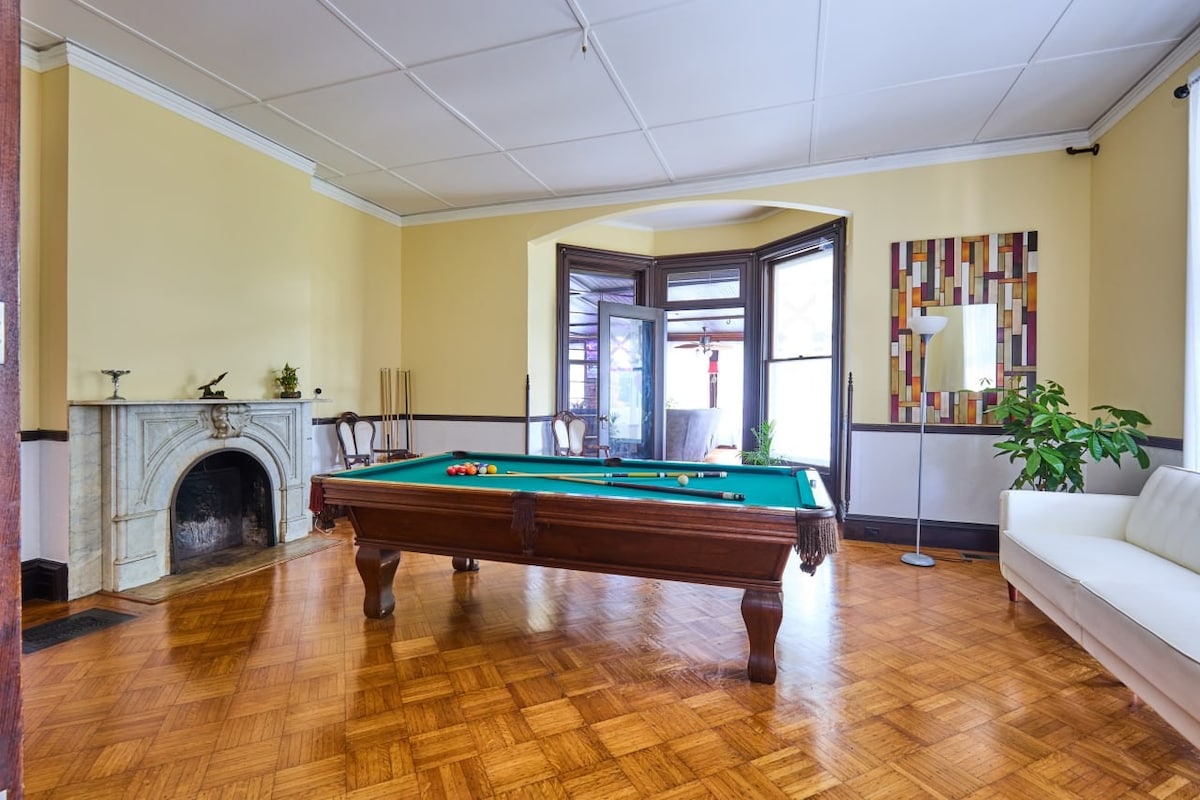 A game room is showcased with a classic pool table at its center, surrounded by warm wood flooring. Natural light enters through large windows, highlighting the room's neutral yellow walls. A cozy seating area is provided by a white sofa, and a decorative plant adds a touch of greenery.