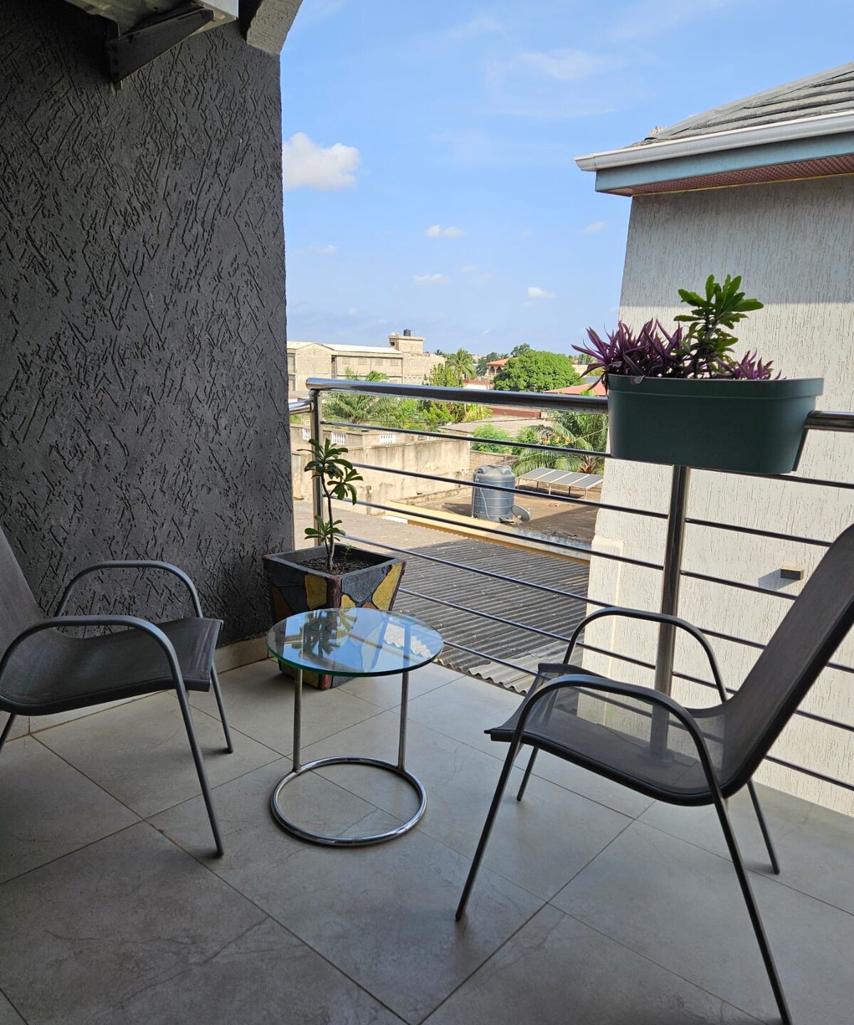 A small balcony features two metal chairs and a glass table. A potted plant adds a touch of green to the space. The railing offers a view of the surroundings, including rooftops and trees under a clear blue sky.