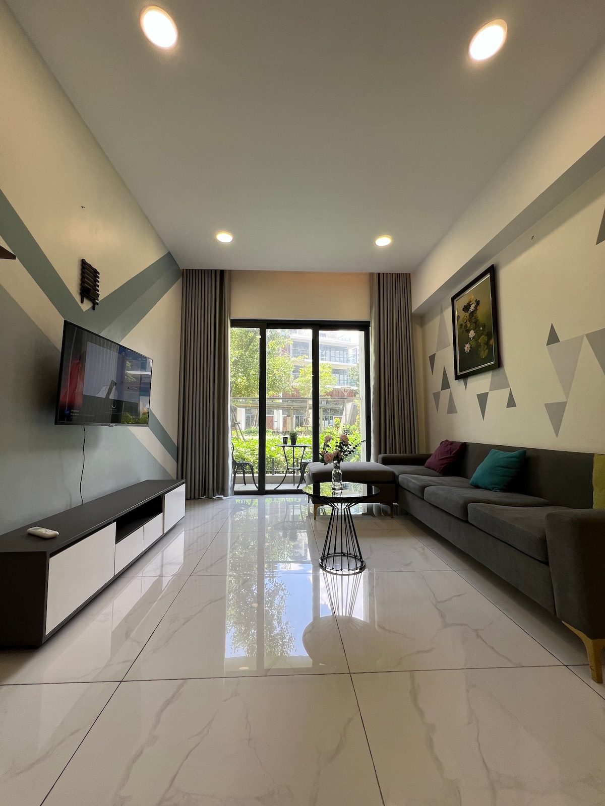 A modern living area features a comfortable gray sofa with colorful cushions, complemented by a sleek coffee table. Large glass doors provide natural light and a view of the greenery outside. A television is mounted on the wall, enhancing the space's convenience.
