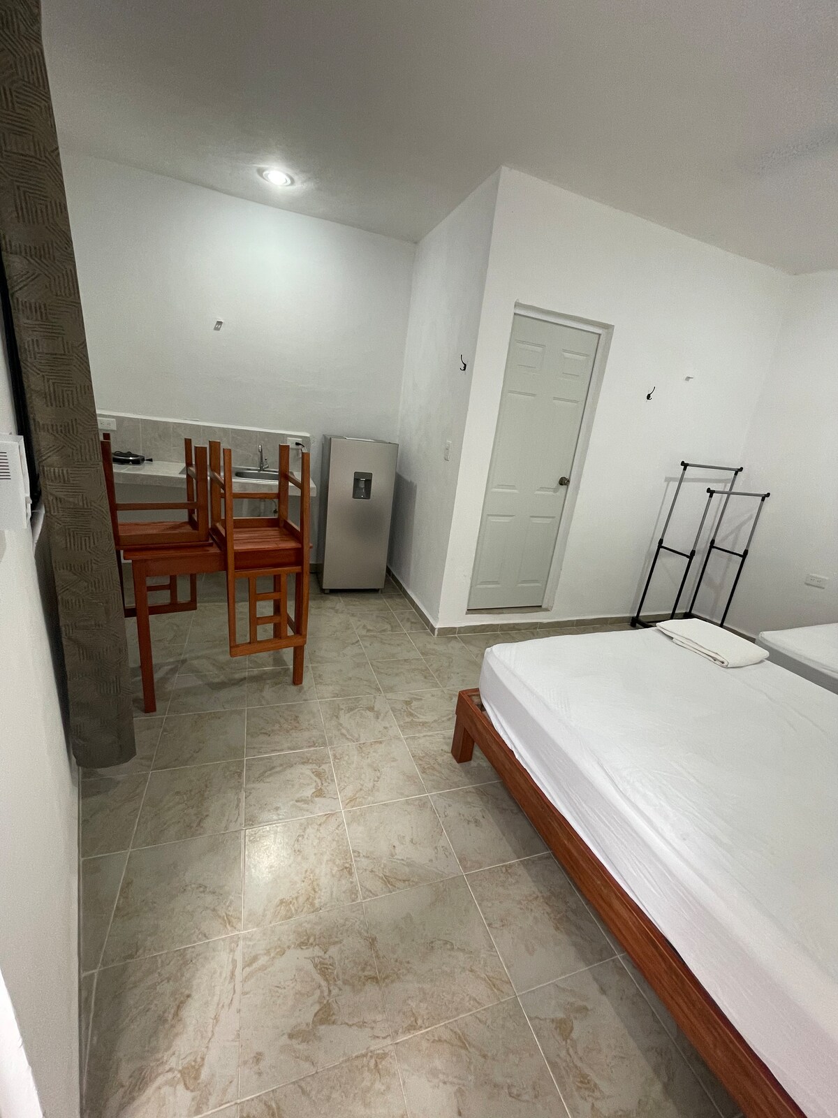 A well-lit room features a spacious bed with light bedding, accompanied by a small refrigerator. Two wooden chairs and a table are arranged near the window. The room showcases a tiled floor and neutral-colored walls, creating a clean and open atmosphere.
