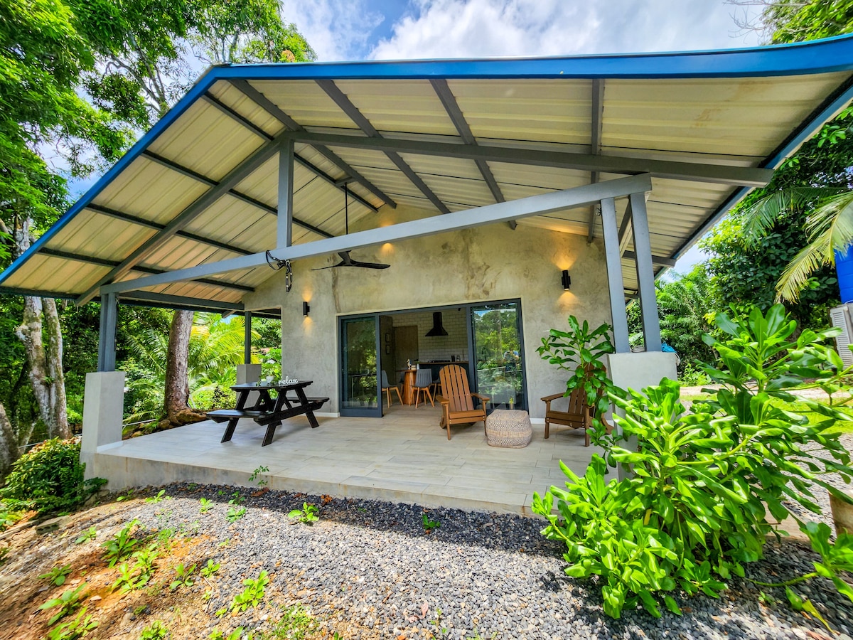 The spacious outdoor patio features comfortable wooden seating arrangements, including two lounge chairs and a picnic table. Lush greenery surrounds the area, complemented by modern lighting fixtures attached to the structure. Large glass doors offer a view of the interior space, blending outdoor and indoor living.