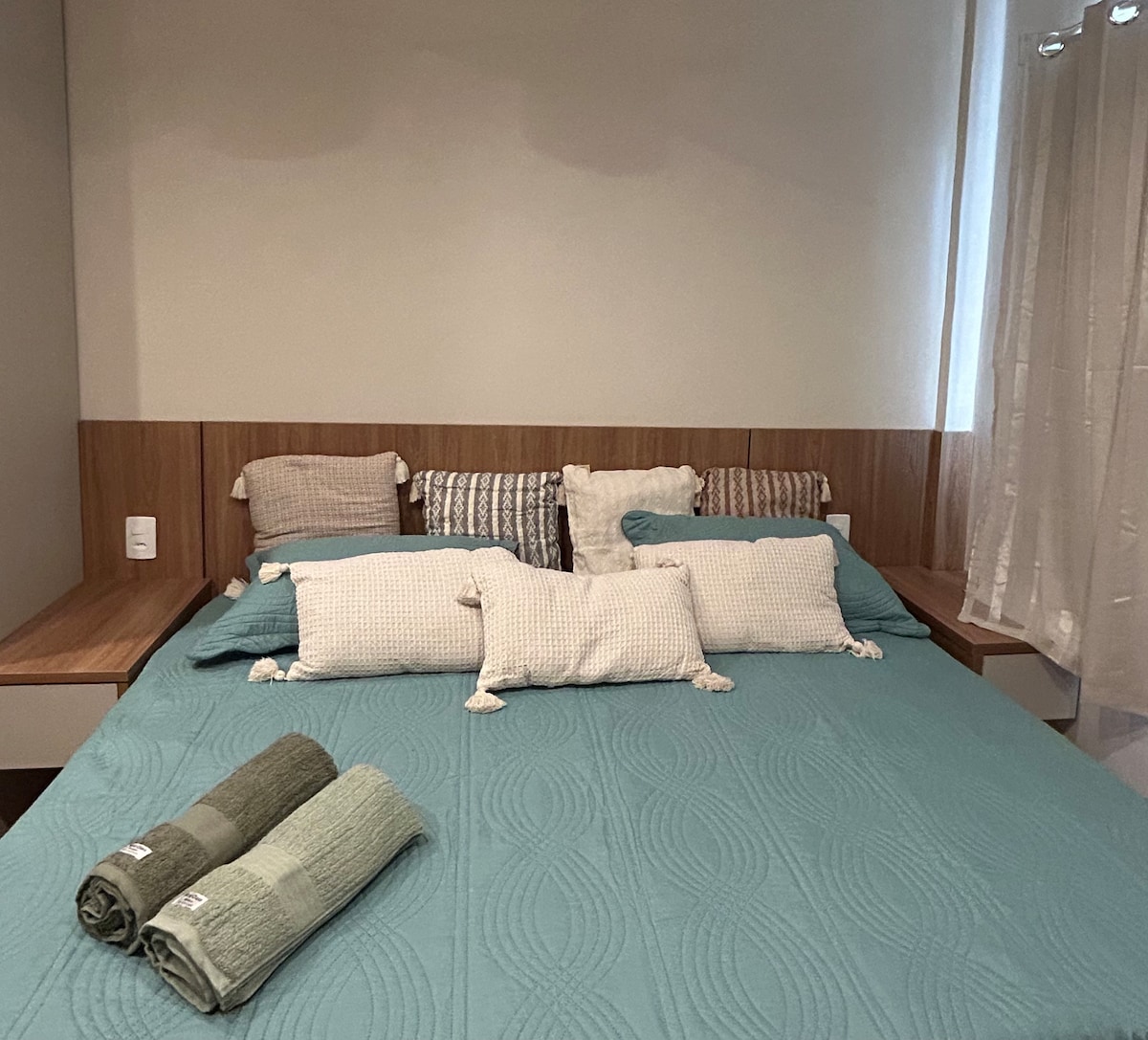 A spacious bed with a light blue quilted cover is adorned with decorative cushions in various patterns. Neatly stacked towels in muted green tones are placed at the foot of the bed. Natural light filters through a nearby window covered by light curtains.