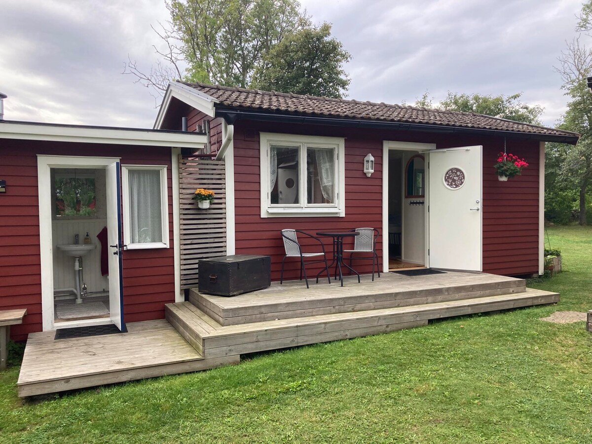 The exterior of a charming red cabin features a wooden deck with a small table and two chairs. A white door with a lantern and decorative elements welcomes guests. Surrounding greenery enhances the tranquil setting, creating an inviting approach to the entrance.