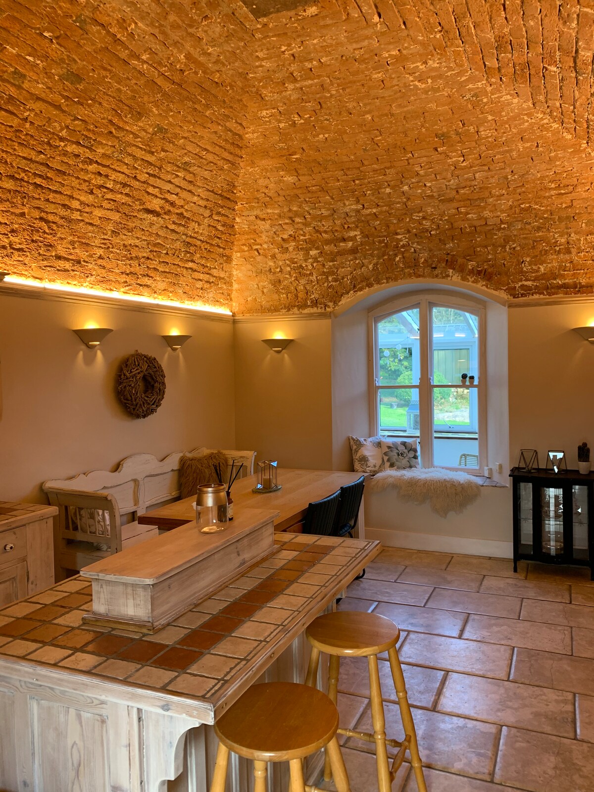 An inviting interior space showcases a warm, brick-vaulted ceiling. A wooden bar with a tiled counter is accompanied by two stools. Soft lighting is provided by wall sconces, and a large window allows natural light to illuminate the area, with greenery visible outside.
