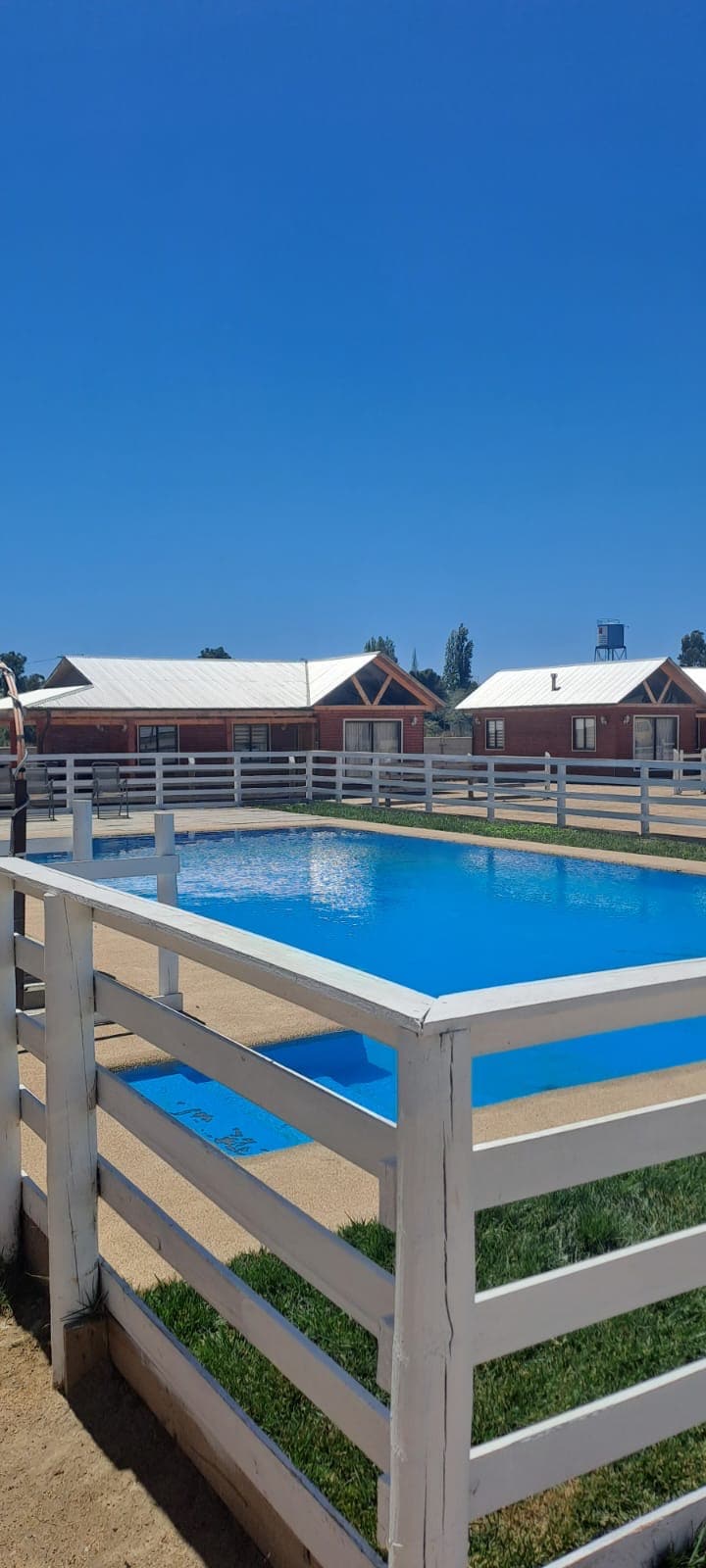 A bright blue pool is surrounded by a white fenced area, reflecting the clear sky. Several rustic wooden cabins are positioned in the background, featuring peaked roofs and light metal accents. A grassy area is visible near the pool, creating a peaceful outdoor setting.