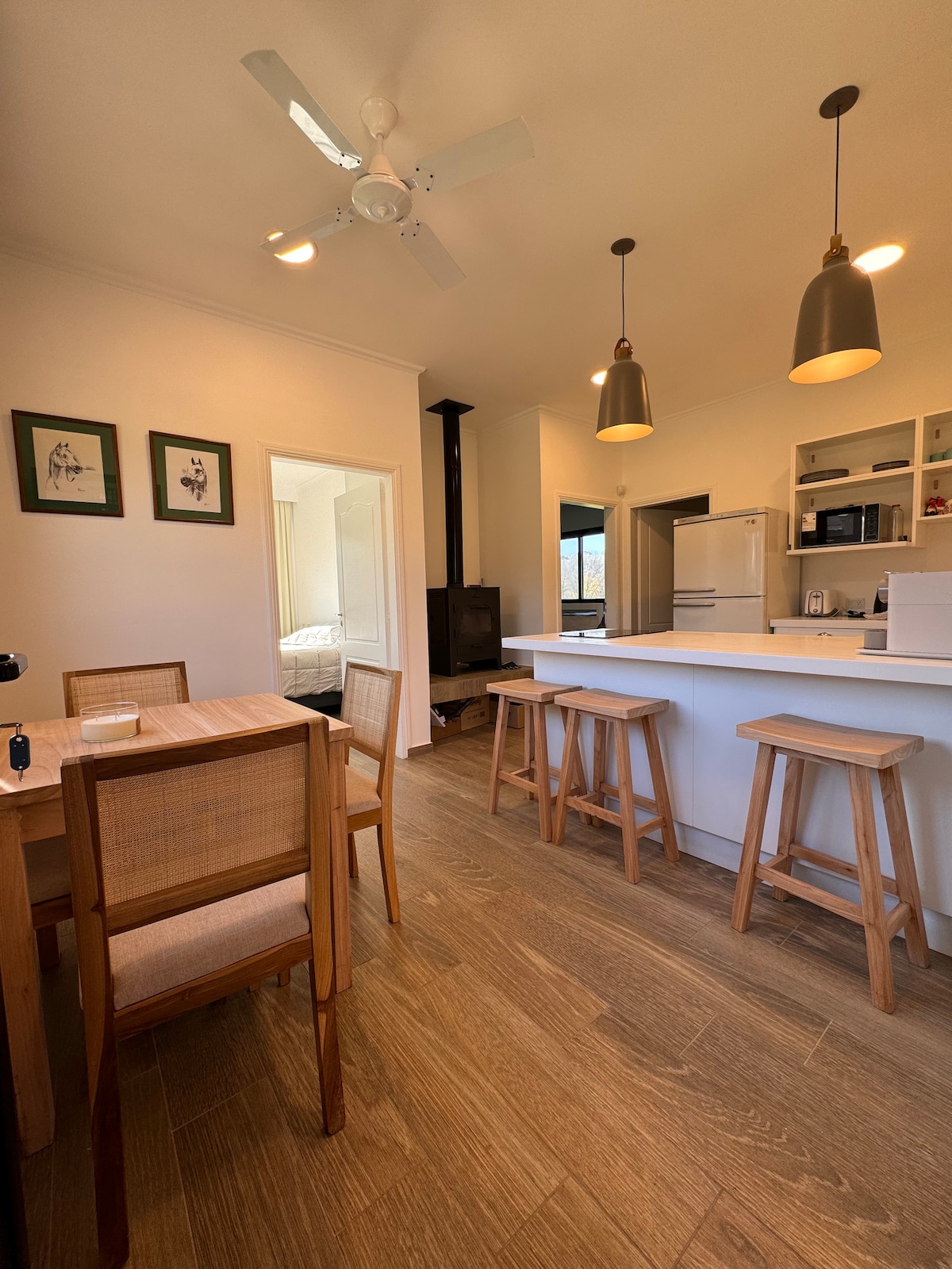 A light and airy kitchen area features a wooden dining table with four chairs and three tall stools at a breakfast bar. Stainless steel appliances are visible, along with open shelving displaying kitchenware. A ceiling fan and modern lighting fixtures are present, enhancing the space's brightness.