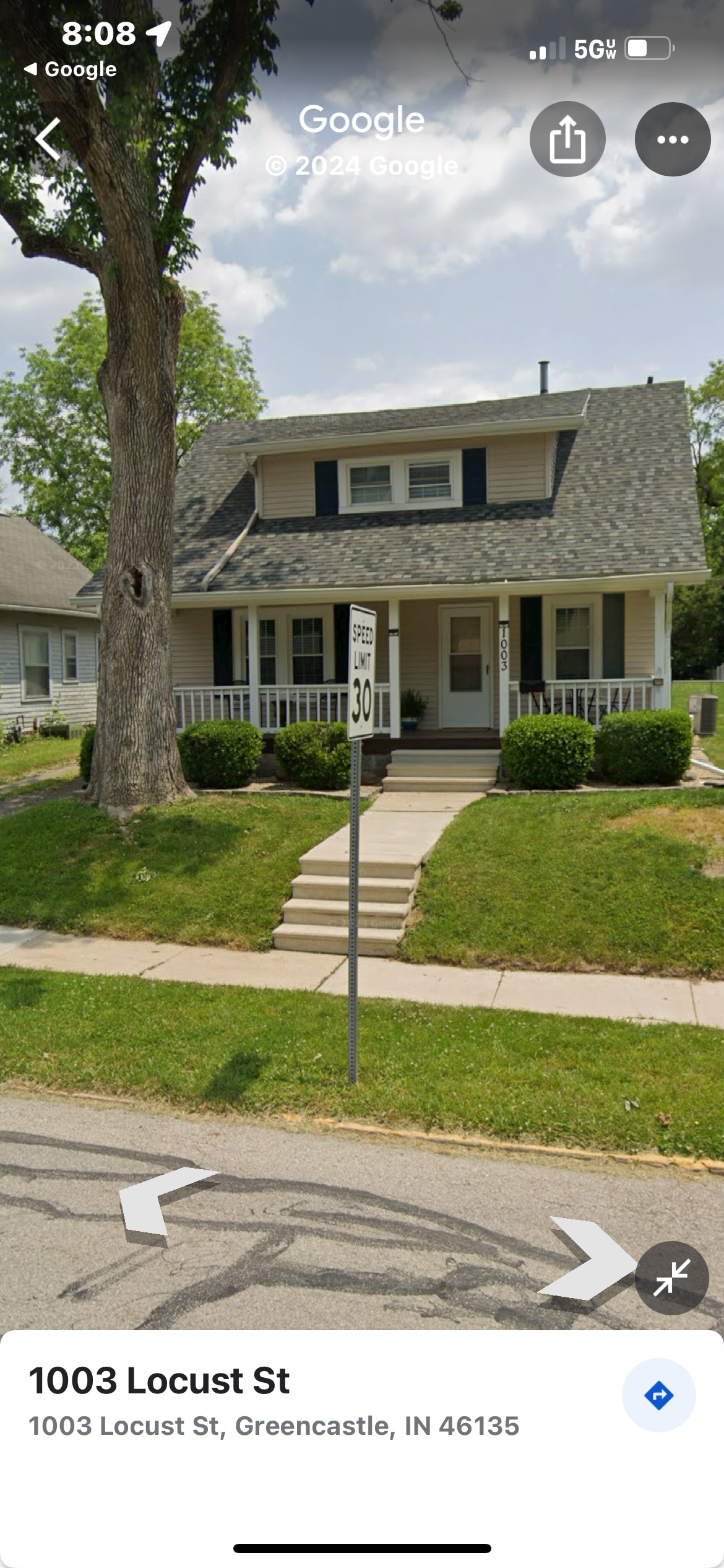 A two-story house with a sloped roof and front porch is framed by symmetrical bushes on either side. A sidewalk leads up to the porch, which features steps and a railing. Grass lines the front yard, and a street sign stands nearby.