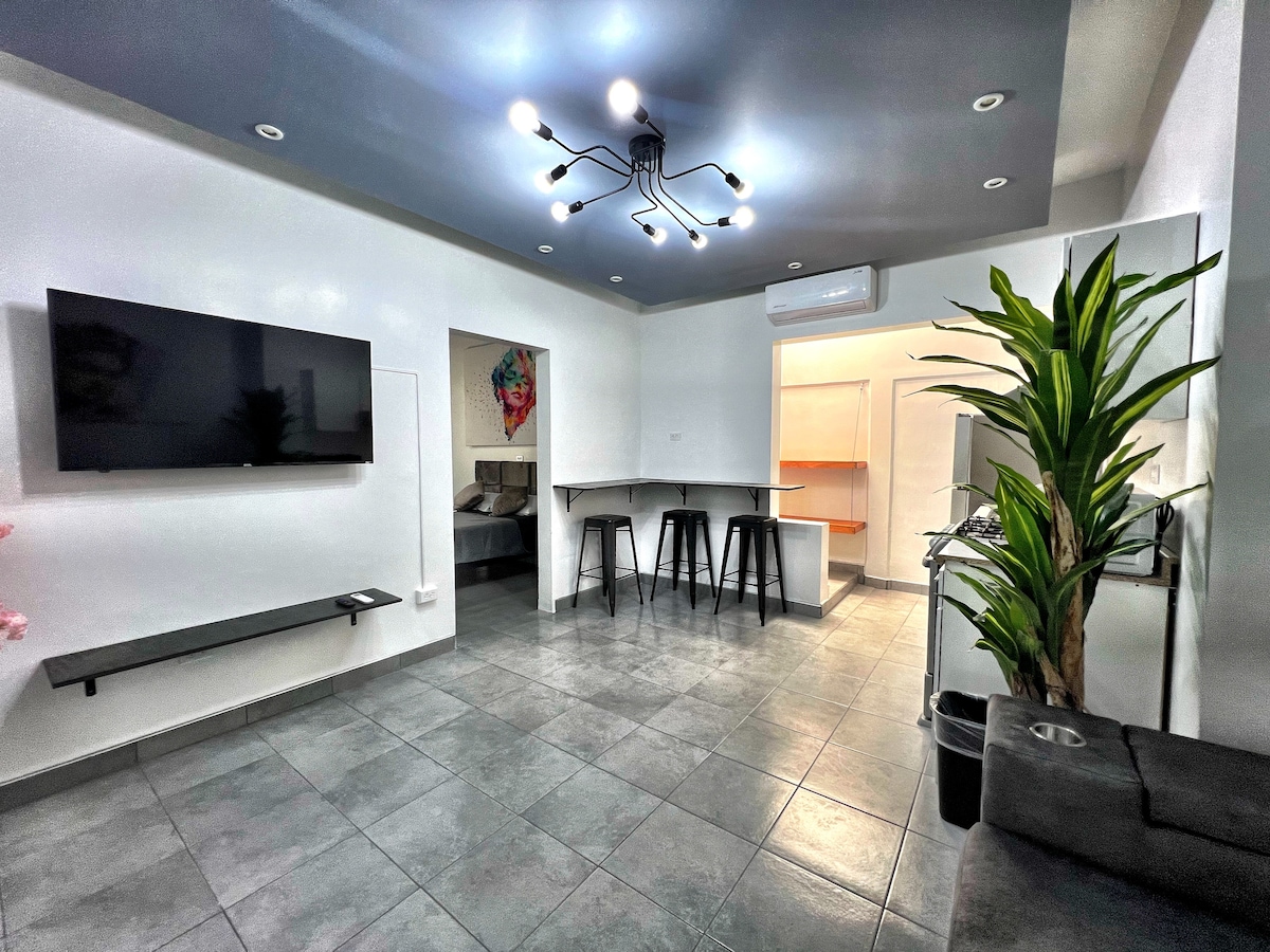 An open-concept living area features modern lighting and a large flat-screen television mounted on a white wall. A bar with high stools separates the kitchen from the lounge, while a potted plant adds a touch of greenery. Soft grey tiles cover the floor.