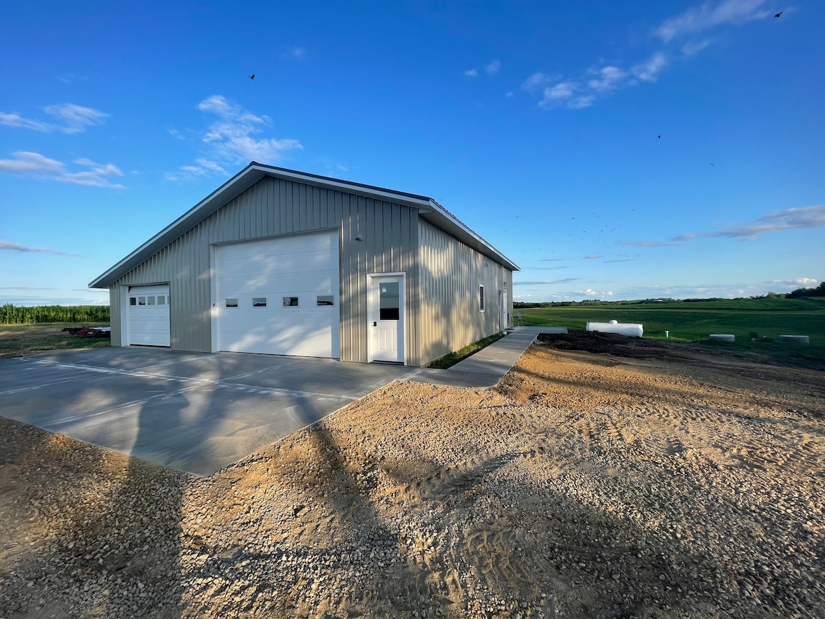 A modern metal building features a large garage door and an entrance door, situated on a gravel surface. Expansive fields are visible in the background under a clear blue sky, with shadows of nearby trees casting patterns on the ground.